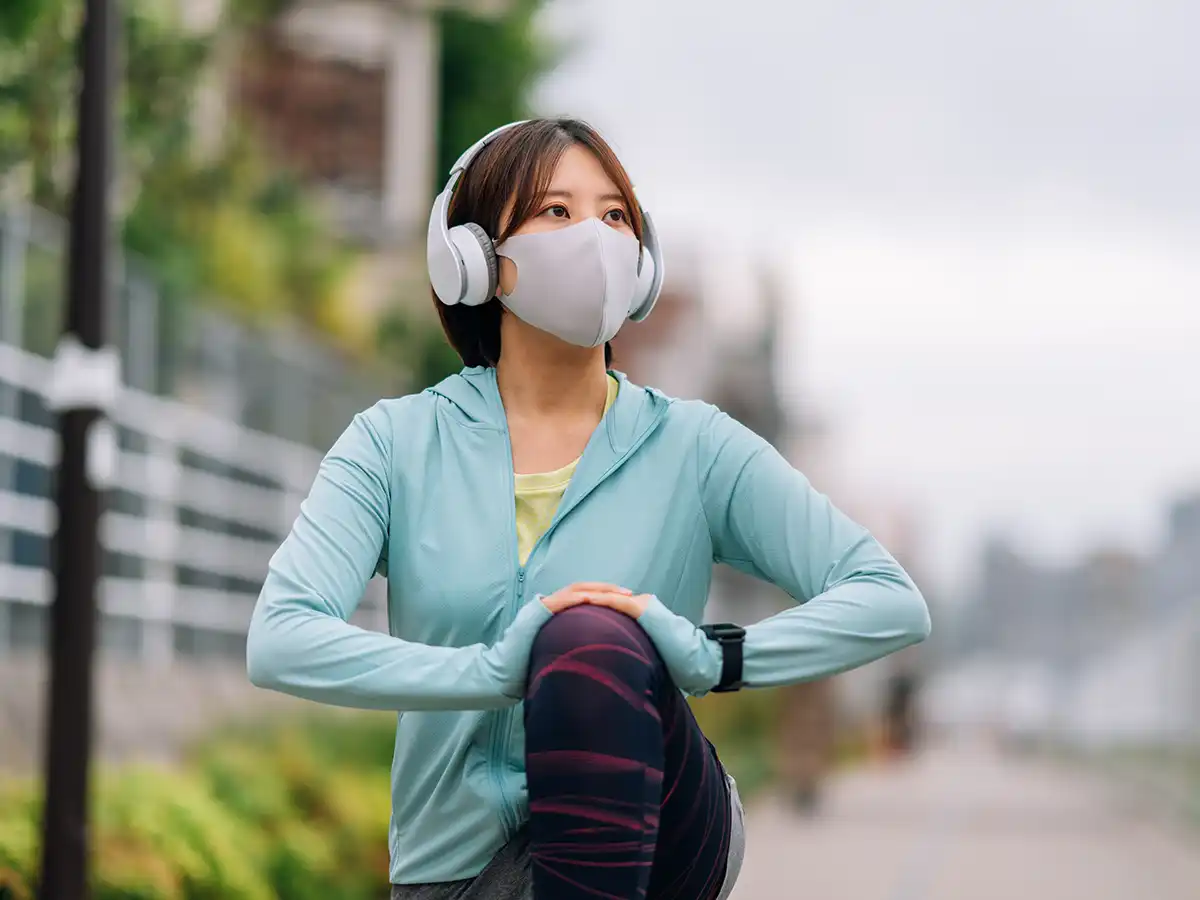 A young Asian woman stretches in a lunge position outside on a running trail. She wears a mask and headphones.